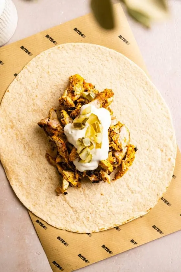 A wrap being built on kraft paper with chicken, sauce and herbs